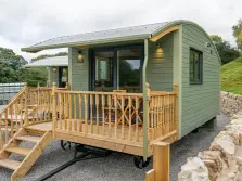 This picture shows a green painted hut on wheels, in front of the hut is a raw timber deck and balustrade around it and steps leading down to a gravel area. Surrounding  the lake district glamping hut are dry stone wall and there are green fields beyond. 