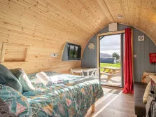This image shows the inside of a camping pod near windermere, through the open door we can see a picnic table, a field, some trees and grass. Inside the pod we can see a wooden ceiling, a bed, a tv and a couch