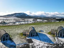 This image shows an aerial view of three lake district glamping pods in the south lakes, it is winter and there is snow on the ground, the shadows are long as it is evening, there is a lake district hill in the background