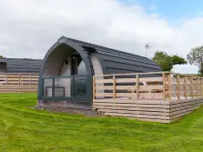 This image shows a lake district glamping pod in a field near Penrith. There is a raised deck with a safety fence around it attached to the pod. In the background another pod can just be glimpsed.