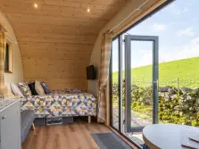 This image shows the inside of a pod for glamping in the lake district. Inside we see a wooden ceiling, a bed and a round table. Through the front glass wall and door we see a traditional dry stone wall and a Lakeland hillside