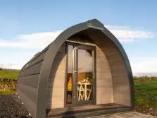 This image shows a modern lake district glamping pod near windermere set in a grassy field, the glass door is partly ajar and we can just see a table and chairs inside.