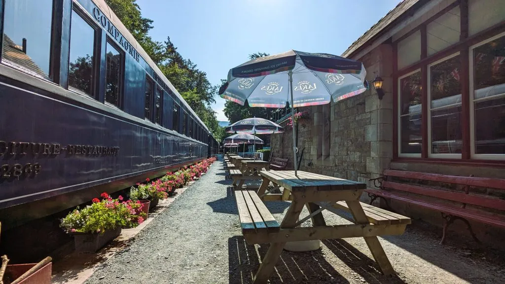 Image of Bassenthwaite Lake Station. photo shows the station, the train ands tables with sun umbrellas