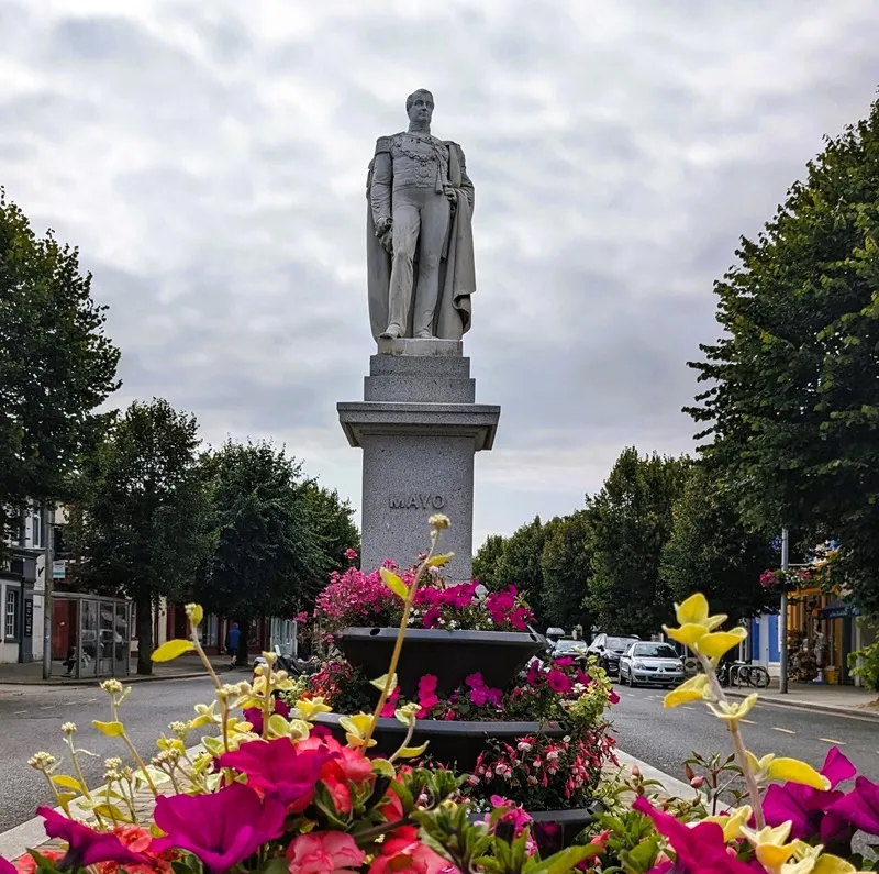 image of Cockermouth Main Street with flower filled planters, trees lining both sides of the street, and the Mayo statue