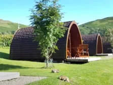 Image of two glamping pods near Keswick, the lake district fells are seen in the background and there is some garden furniture visible in front of one of the pods and there is a small tree in the foreground