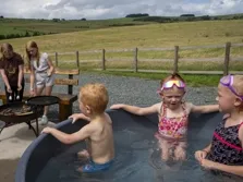 Image of three children in a hot tub, behind them their parents are cooking on a bbq outside a camping pod. There are fields, rural lake district views and trees in the background under a summer sky