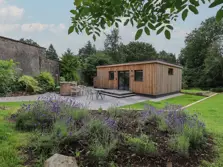 Here we see a picture of a modern, very square glamping cabin set on gravel and surrounded with grass. The cabin has two windows and a door and outside it there is a hot tub and garden furniture.to one side of the cabin there is a farm building and behind there are trees. There is a raised rocky bed of lavender bushes in the foreground. Eastern lake district
