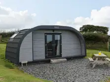 This image show a grey glamping pod in a field, the pod is wider than it is long. There is a gravel area in front of the pod with a picnic table and bench on the gravel. In the background is grass, a hedge and trees. The sky is pale blue over the site that provides glamping in the lake district.