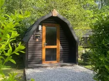This image shows a glamping pod front elevation with gavel area in front. There are tall bushes and trees surrounding the pod on three sides. South Cumbria