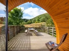 This image shows the view from inside a lake district camping pod, outside we can see a timber deck, a picnic table, green fields and trees under a blue sky near Ullswater
