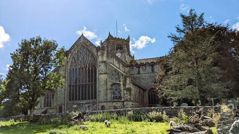 image of the exterior of the Cartmel Priory, the Priory Church of St. Mary and St. Michael, Cartmel in Cumbria. photo shows the big stained glass window from the outside as well as sheep in the fields