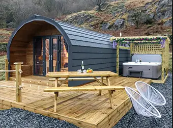 This image shows a long, low, round roofed glamping pod set on a timber deck. There is a wooden picnic table and benches on the deck and, set further back, a hot tub under a pergola decorated with flower garlands. Two modern metal chairs are set beside the deck. In the background we see a rocky hillside with trees on it. This is a glamping pod near Hartsop, Glenridding.