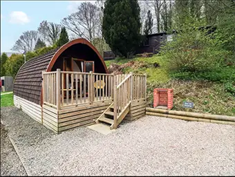 This image shows a round roofed glamping pod near Keswick in the lake district with a raised deck and steps leading down to a gravel area. There is a brick bbq to one side and in the background is a timber cabin and a bank with trees on it. 